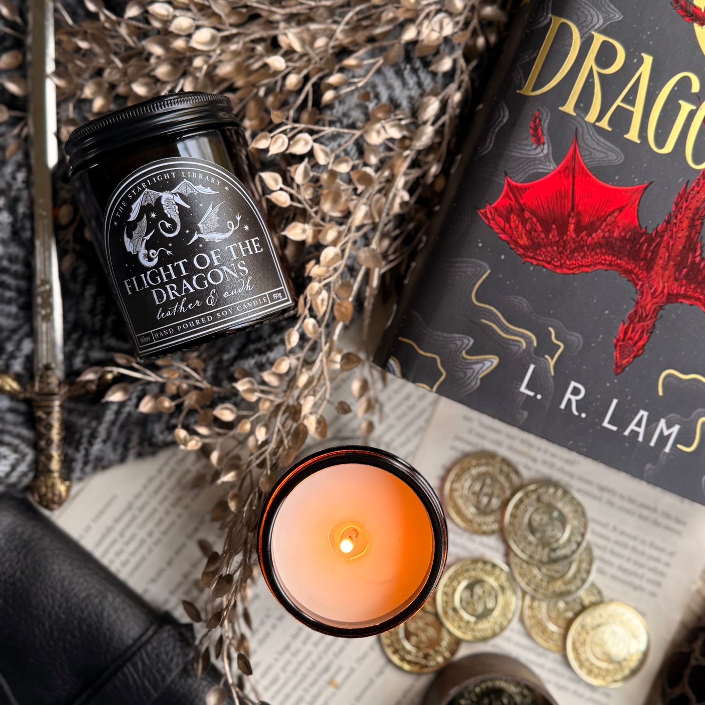 A bookish Flight of the Dragons candle, in an amber jar with a black label, laying down beside a lit candle and a book with a red dragon on the front.