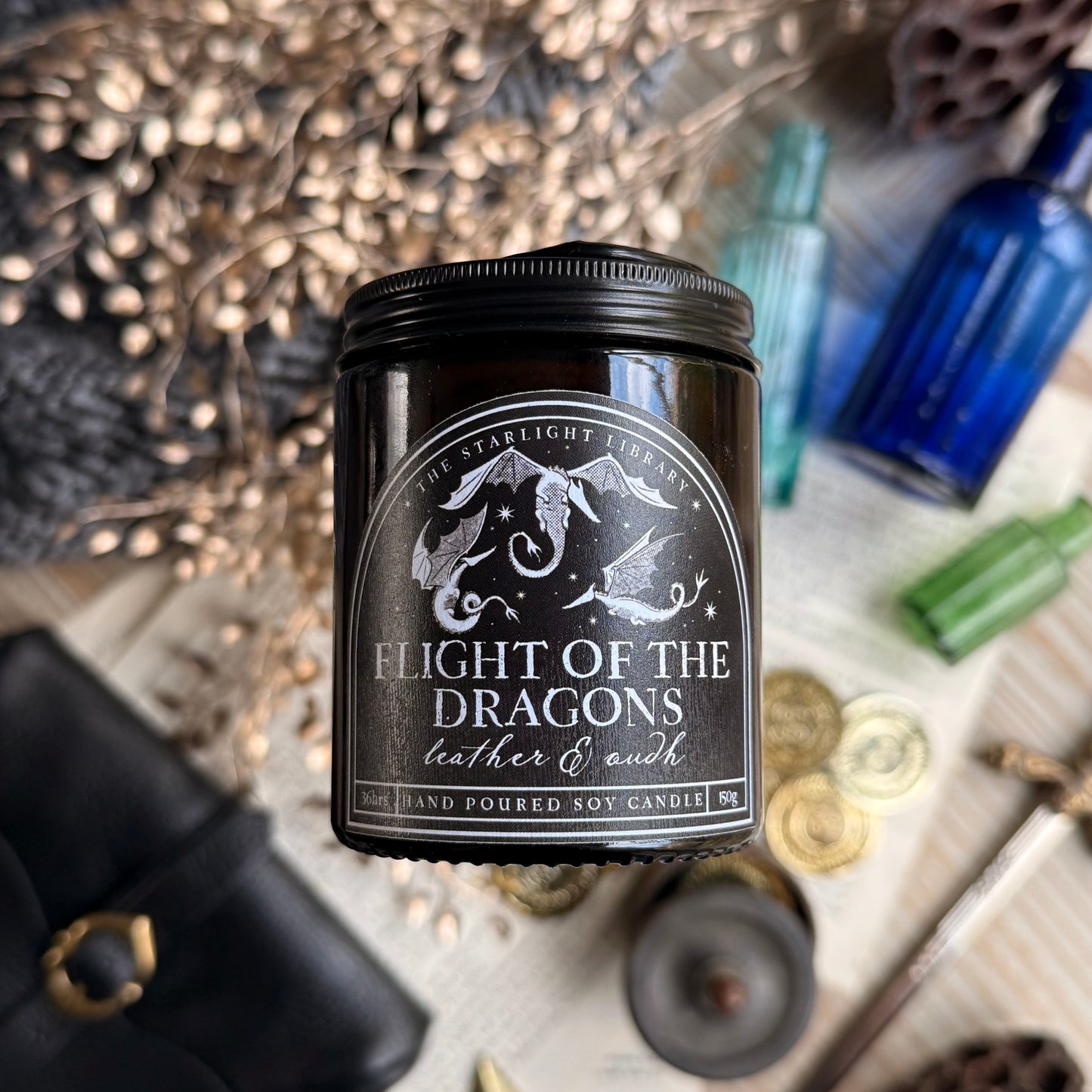 A bookish Flight of the Dragons candle, in an amber jar with a black label, floating above a background of book pages, gold coins and blue glass poison bottles. 