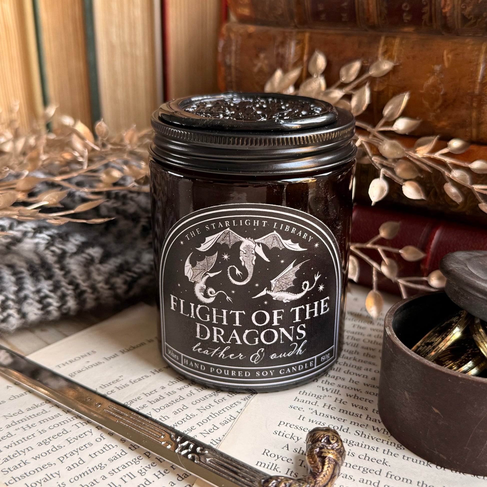 A bookish Flight of the Dragons candle, in an amber jar with a black label, surrounded by old leather books, cosy knits and a miniature sword.