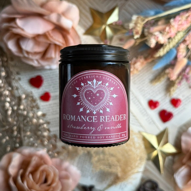 A bookish Romance Reader candle, in an amber jar with a dark pink label, floating above a background of book pages, hearts and dried flowers.