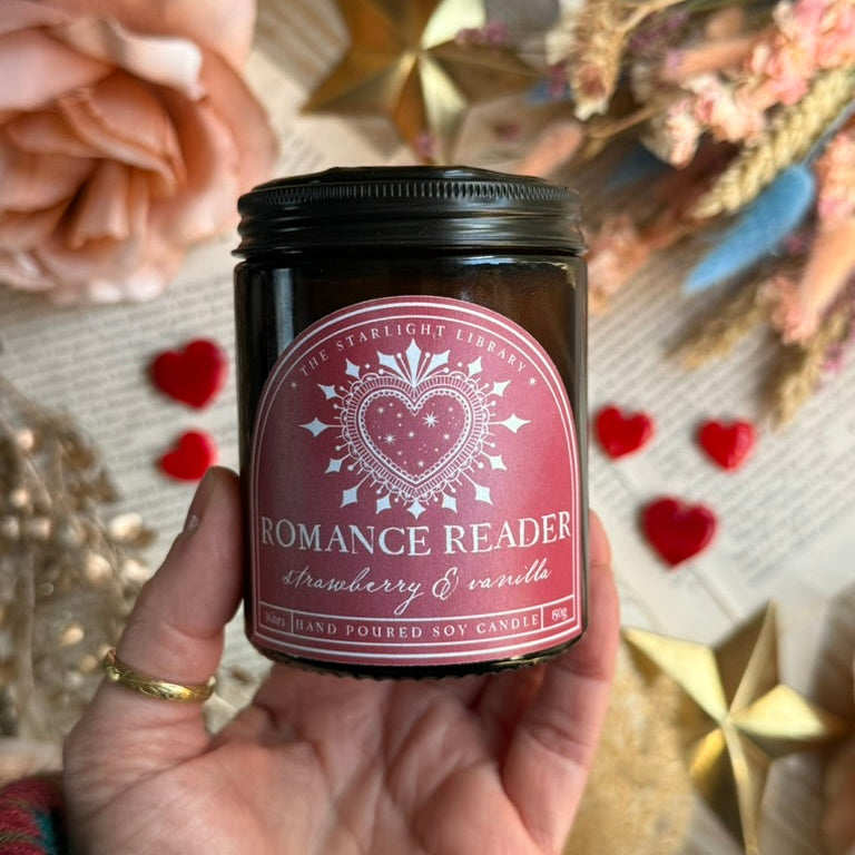 A bookish Romance Reader candle, in an amber jar with a pink label, held in a woman's hand above a background of book pages, hearts and dried flowers.