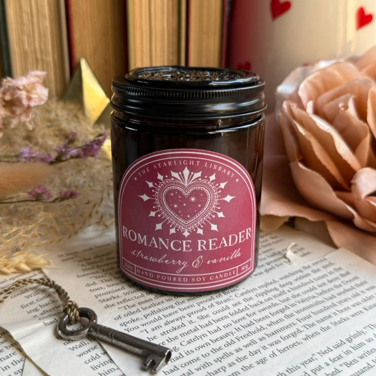 A bookish Romance Reader candle in an amber jar, with a dark pink label and black lid on.