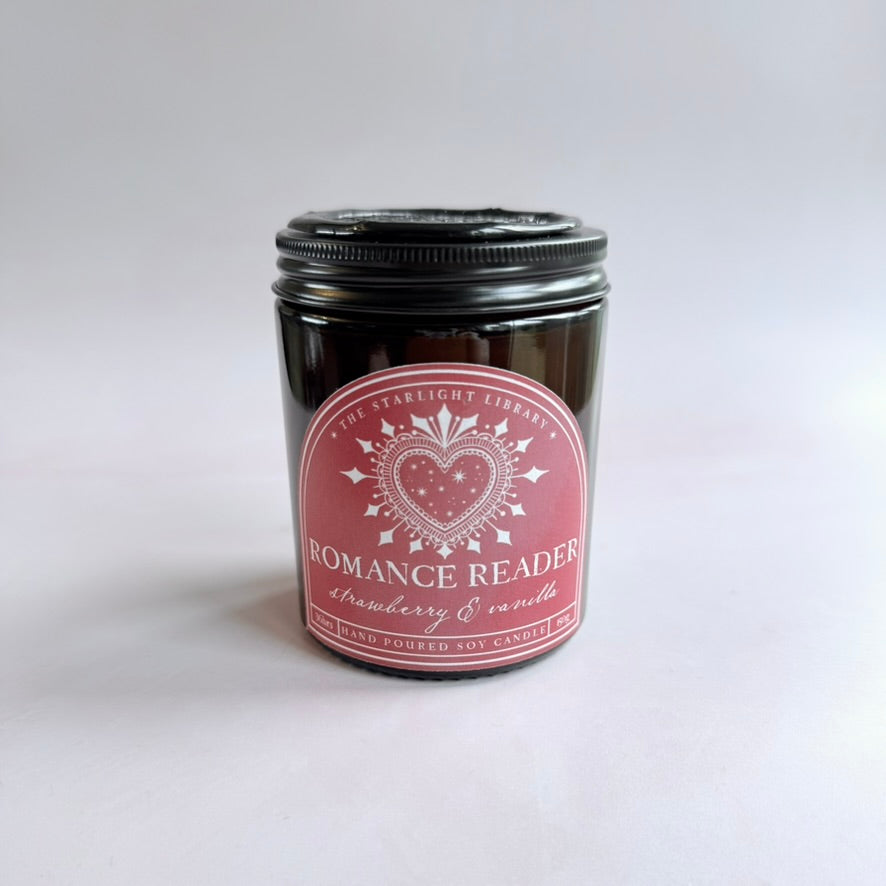 A bookish Romance Reader candle, in an amber jar with a pink label, against white background.